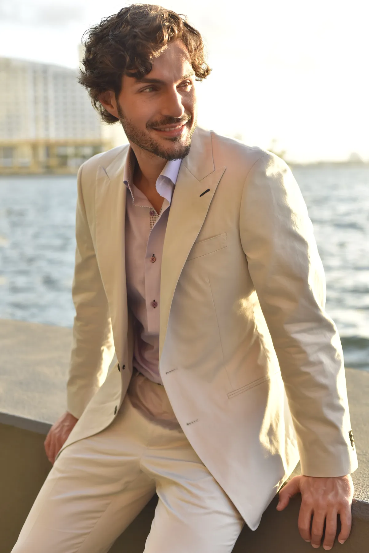 A man wearing a cream linen suit standing along a Miami waterfront at sunset, showcasing lightweight warm weather tailoring.