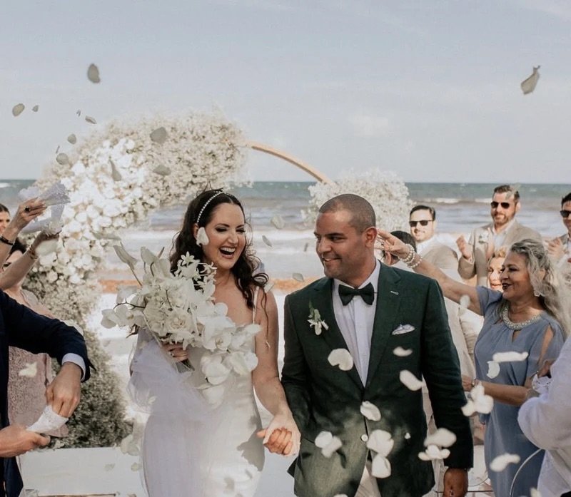 Groom in a forest green suit with black bow tie walking with his bride through flower petals at a beach wedding ceremony