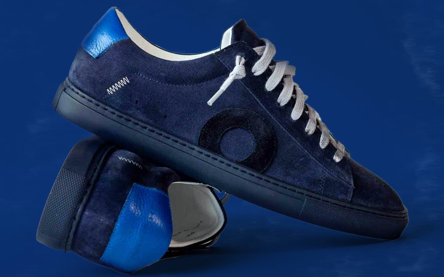 Oliver Cabell Low 1 sneakers in navy suede with metallic blue heel counter and Margom sole, displayed in crossed studio product shot against blue background