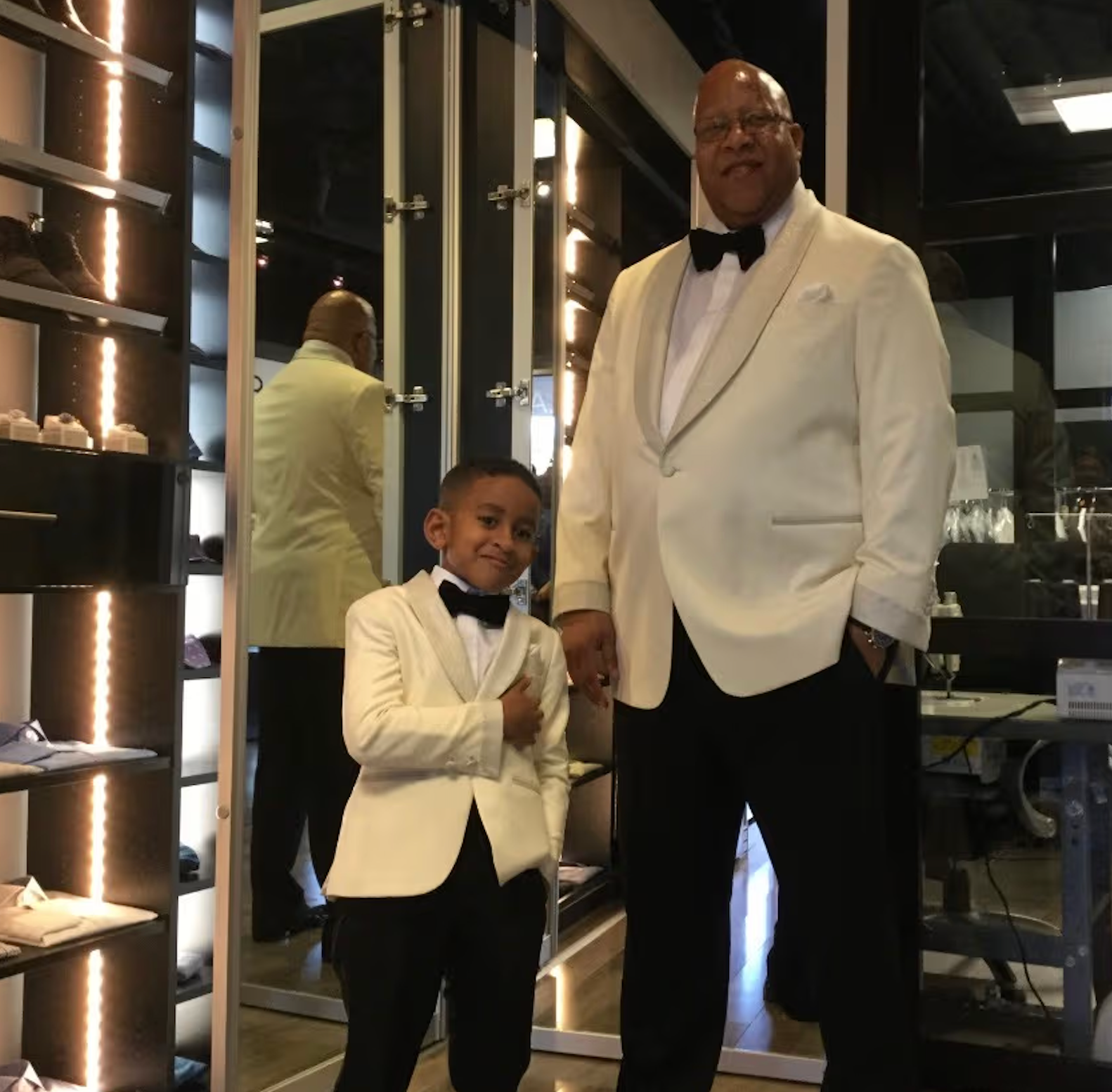 Father and young ring bearer wearing matching ivory white dinner jacket tuxedos with black shawl lapels and bow ties in formalwear shop, showing miniature ring bearer tuxedo styling