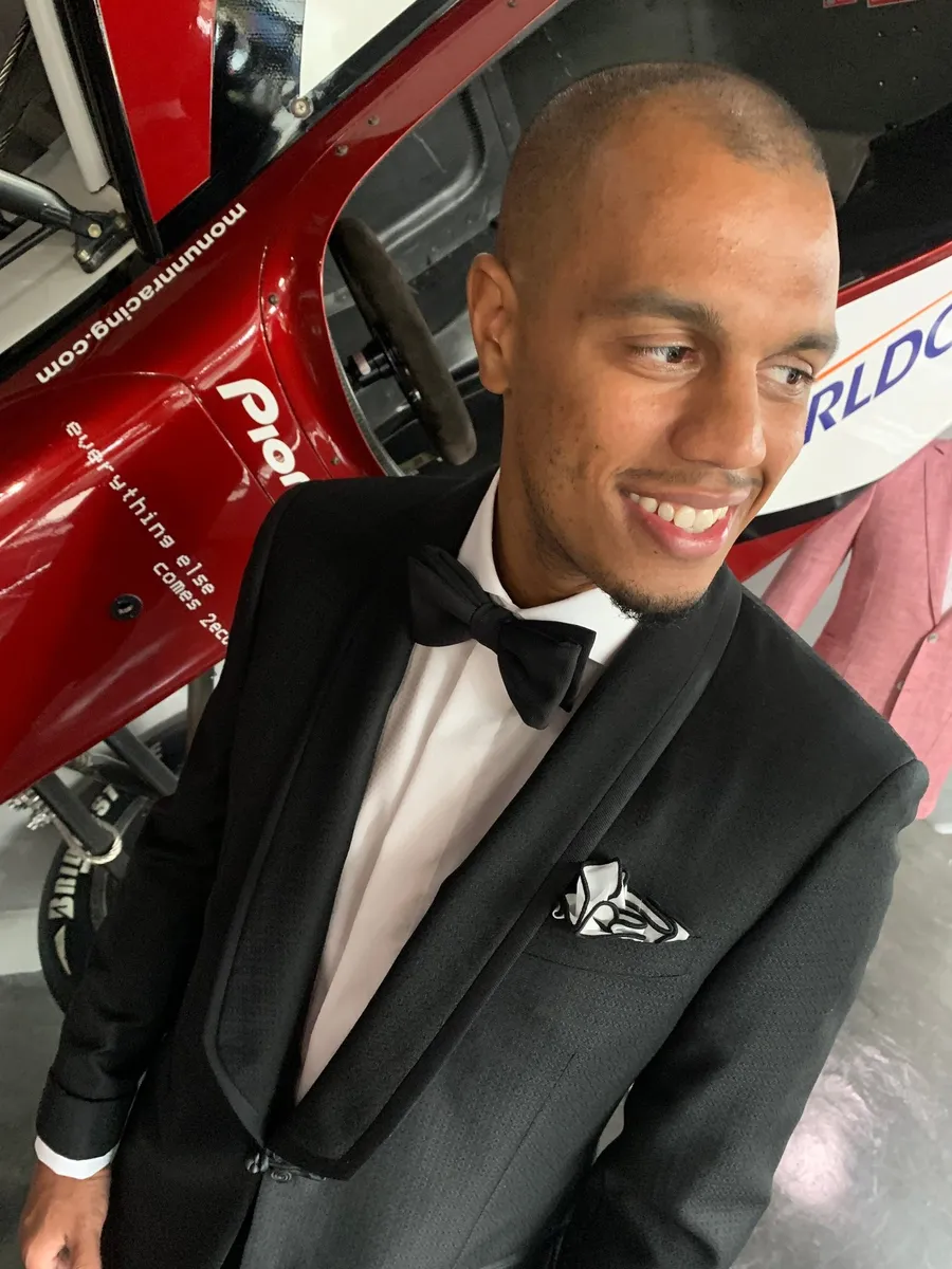 Client in black tuxedo with bow tie posing beside red race car at formal event
