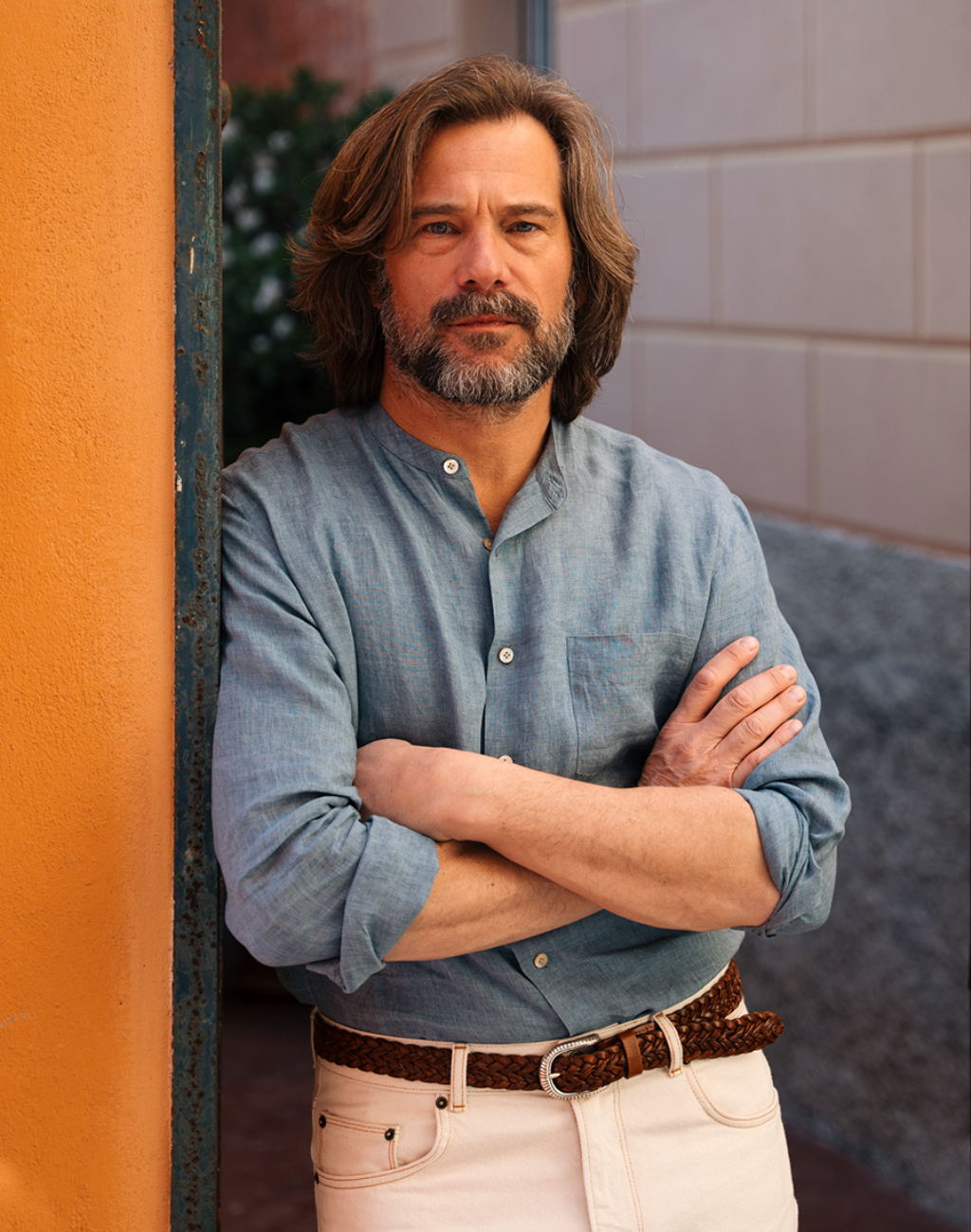 Man wearing a muted blue band-collar shirt with rolled sleeves and pale trousers beside an orange wall