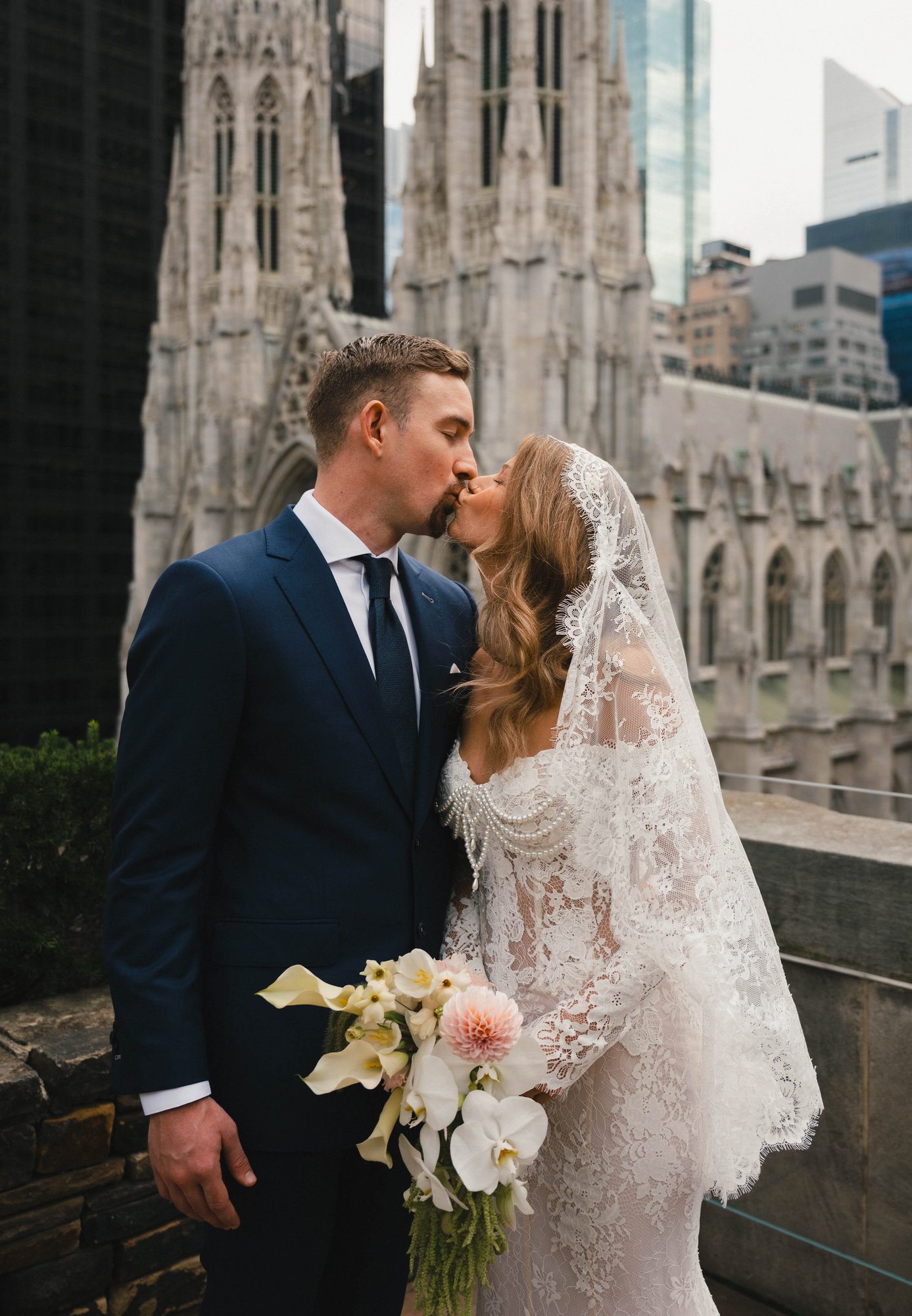 The couple embracing outside a grand building entrance, the custom suit detailing visible in the wedding portraits.