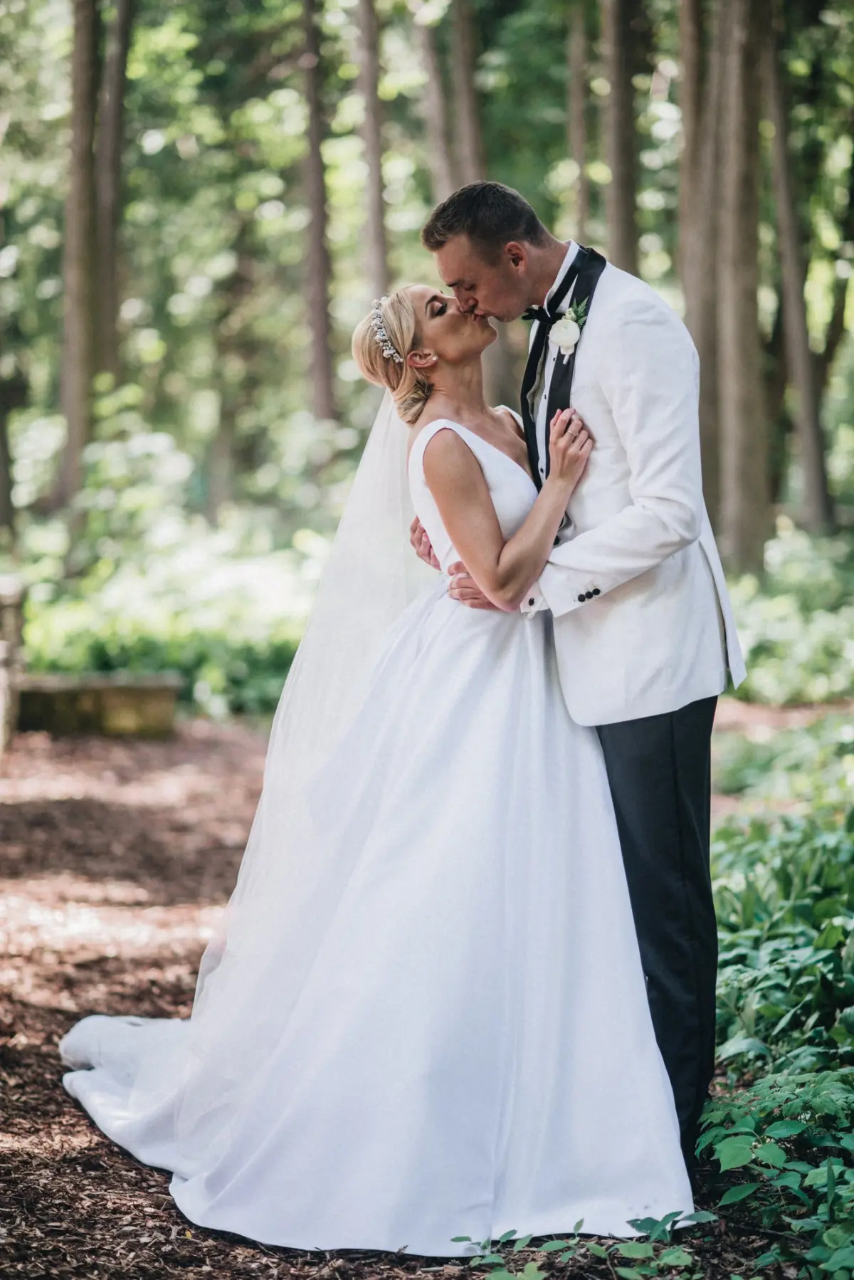 Groom in white dinner jacket with black lapels embracing bride in wooded setting, demonstrating custom fit on a tall frame