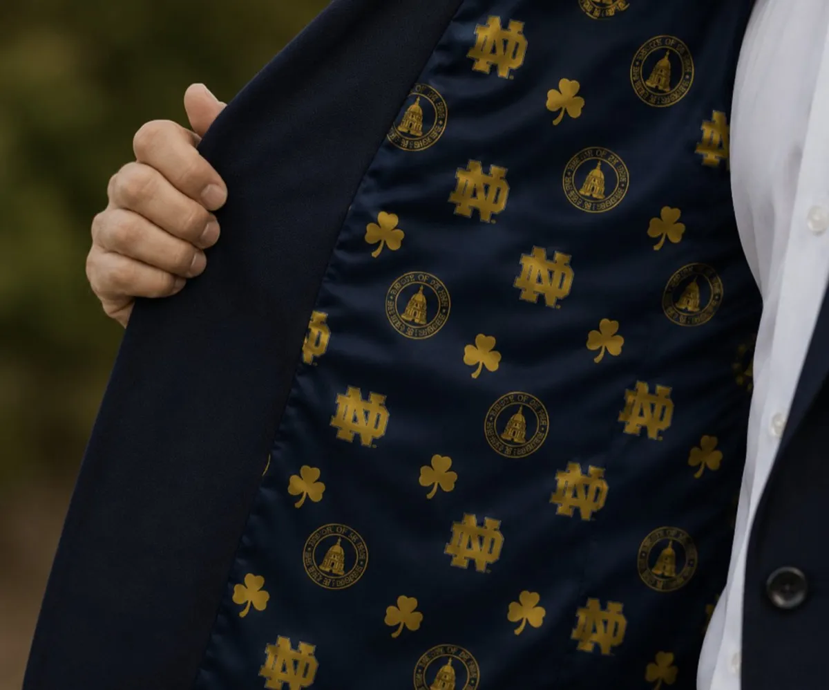 Navy blue suit jacket held open to reveal a gold Notre Dame University lining with interlocking ND monogram, university seal, and shamrock motifs, custom tailored by Bespoke By CB.