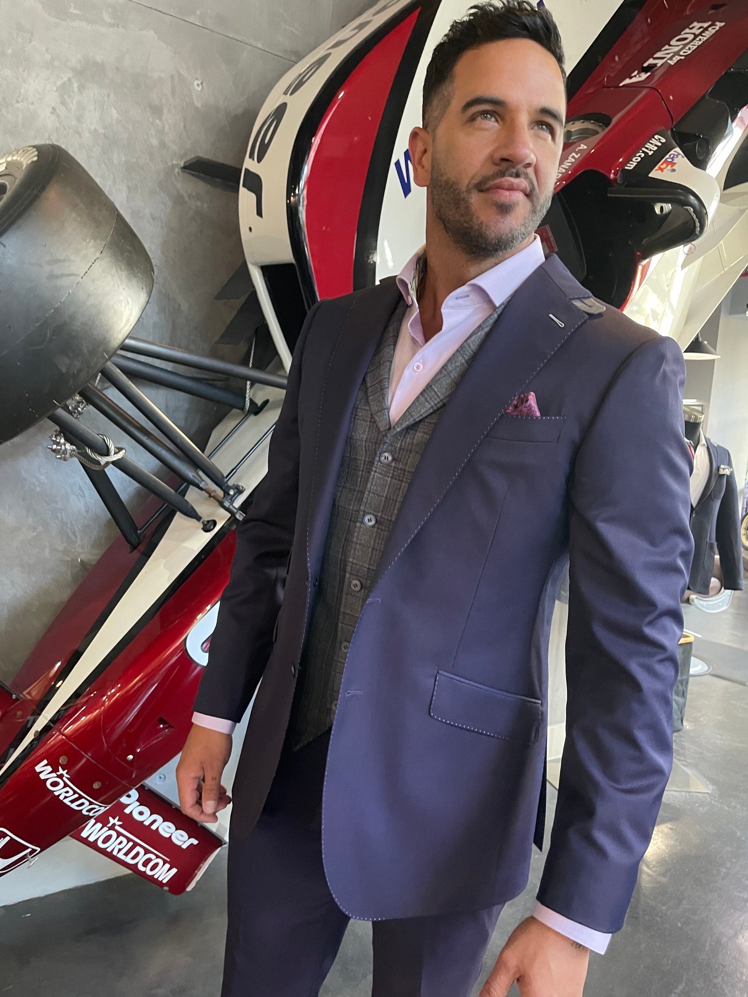 Man in a custom navy suit and plaid vest posing next to a red racing car at Bespoke By CB