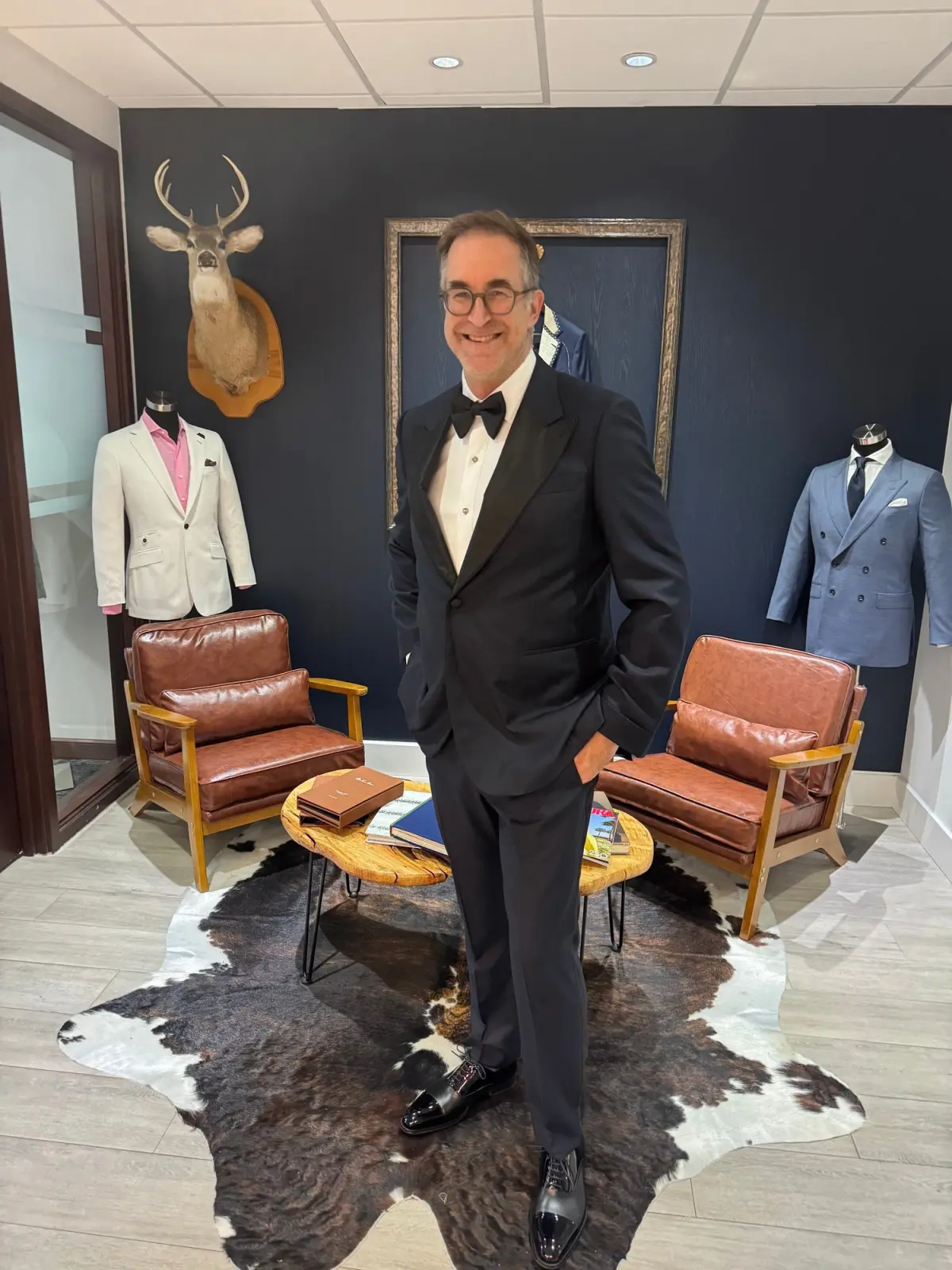 Man wearing a custom black tuxedo in the Bespoke By CB Miami showroom with leather chairs and suit mannequins