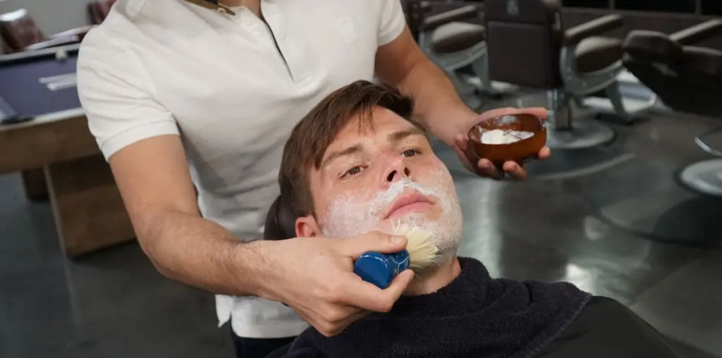 Barber in a white polo shirt lathering a seated client's face with a shaving brush, holding a wooden bowl of shaving cream in a barbershop