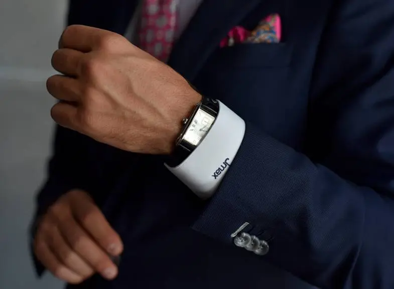 Close-up of a navy suit sleeve pulled back to show a monogrammed white shirt cuff and silver rectangular watch, with a pink tie and paisley pocket square