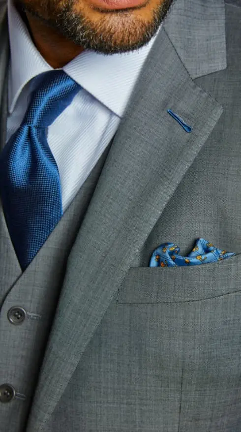 Close-up of a grey three-piece suit with a white shirt, blue textured tie, blue lapel buttonhole and blue patterned pocket square