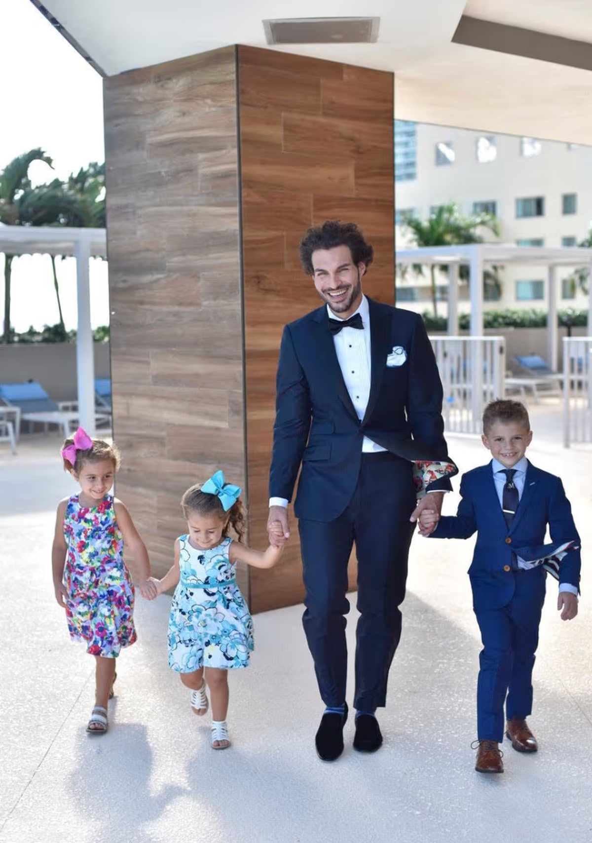 Groom in navy blue wedding tuxedo walking with ring bearer in matching royal blue suit and flower girls at outdoor resort ceremony, showing coordinated wedding party attire