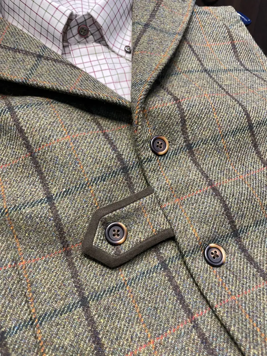 Close-up detail of custom green tweed shooting waistcoat showing windowpane check pattern, horn buttons, hexagonal cartridge pocket with contrasting trim, and notched lapel collar