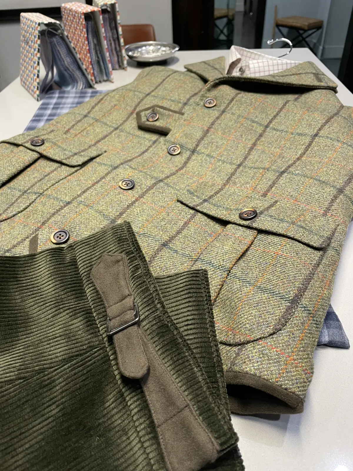 Flat-lay of custom olive green tweed shooting vest with five-button front, hexagonal cartridge pocket, flapped bellows pockets, and horn buttons, paired with olive corduroy trousers on tailor's cutting table