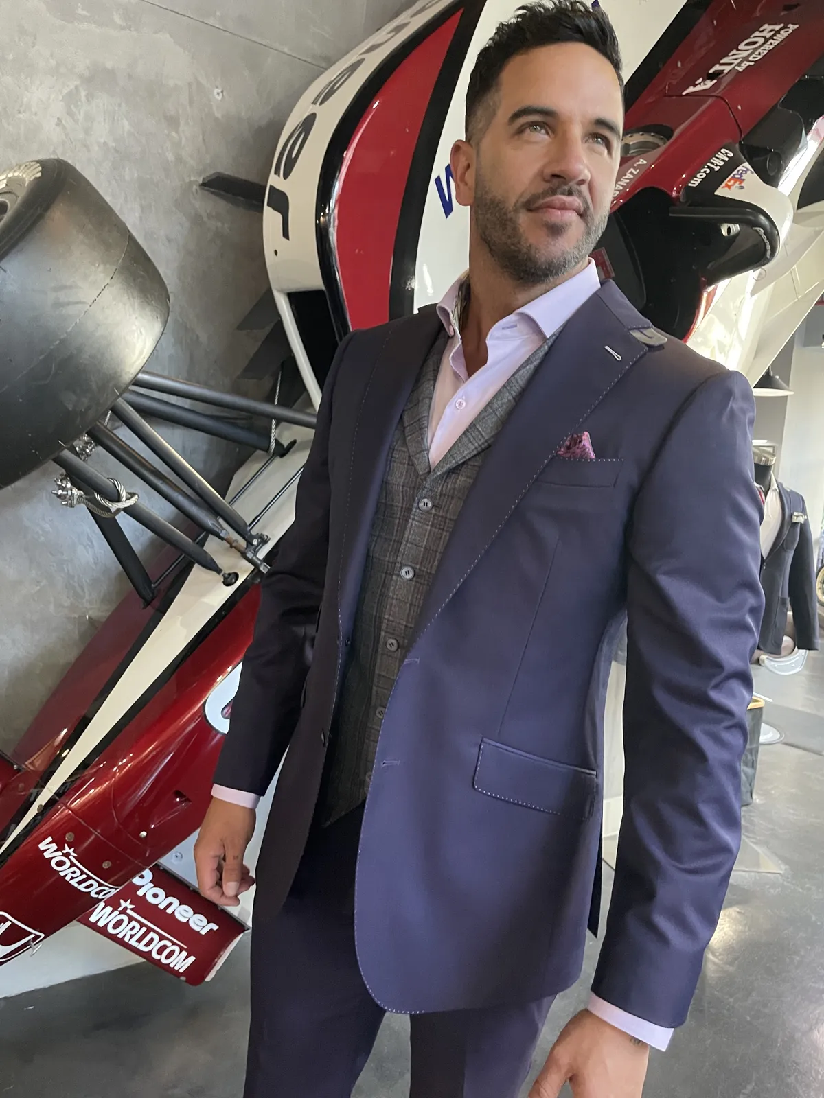 Man wearing a dark navy jacket and trousers with a grey plaid waistcoat, pale pink open-collar shirt and pink pocket square beside a race car