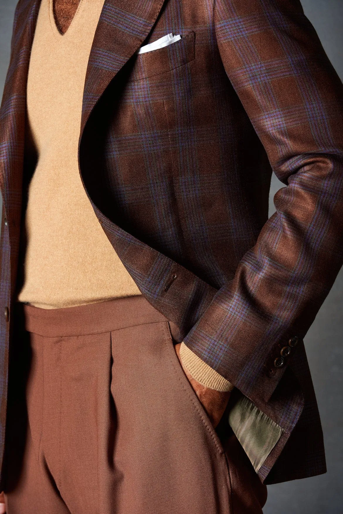 Close-up of a brown and blue plaid blazer over a camel V-neck sweater with rust-colored trousers and a white pocket square