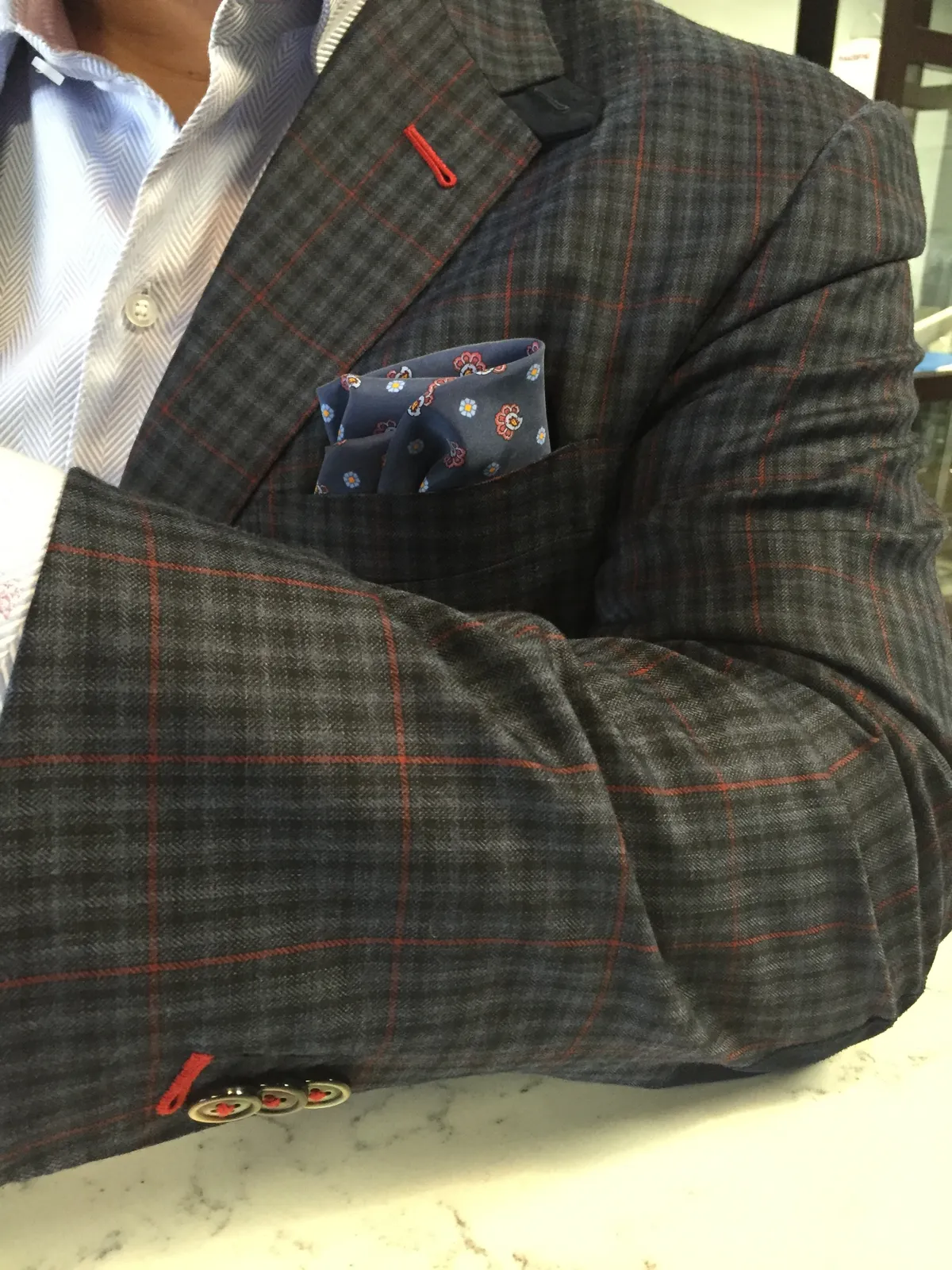Close-up of a grey checked sport coat with red windowpane lines, red buttonhole stitching and a navy floral pocket square over a light blue striped shirt