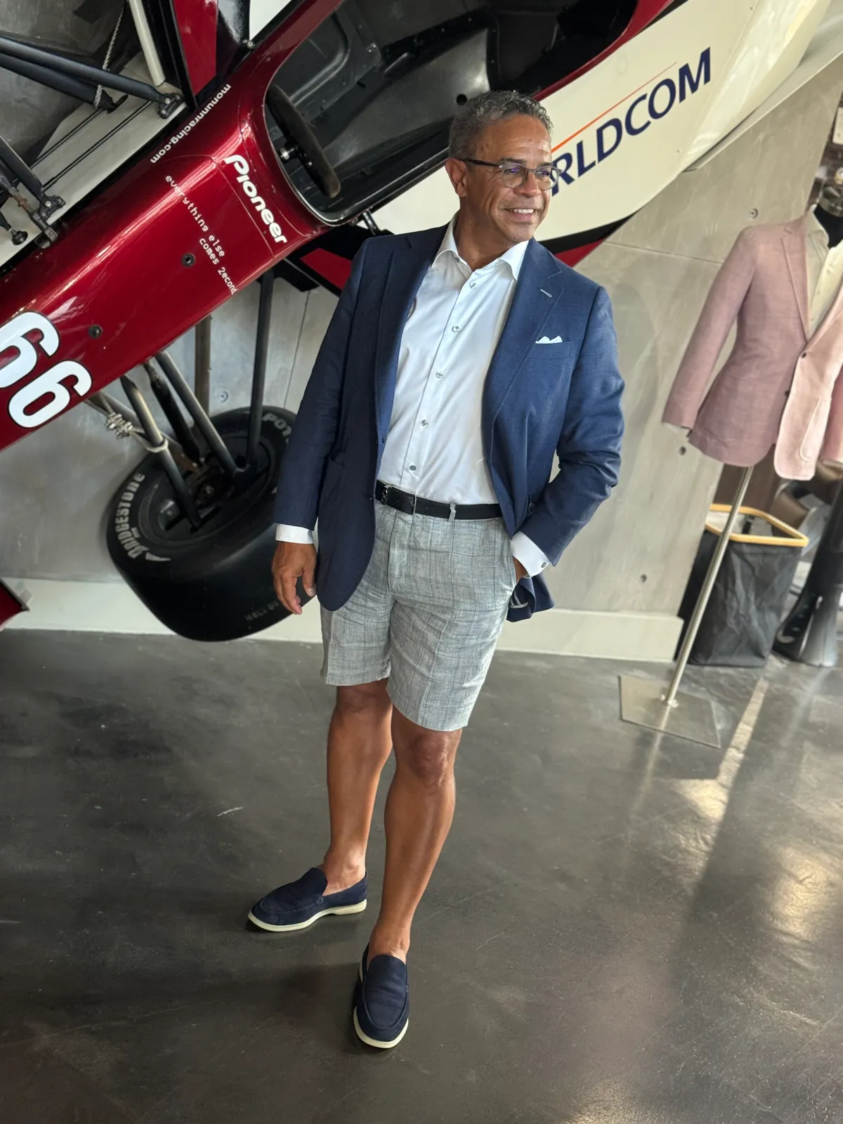 Man in a navy blazer and grey tailored shorts with loafers at an aircraft hangar.