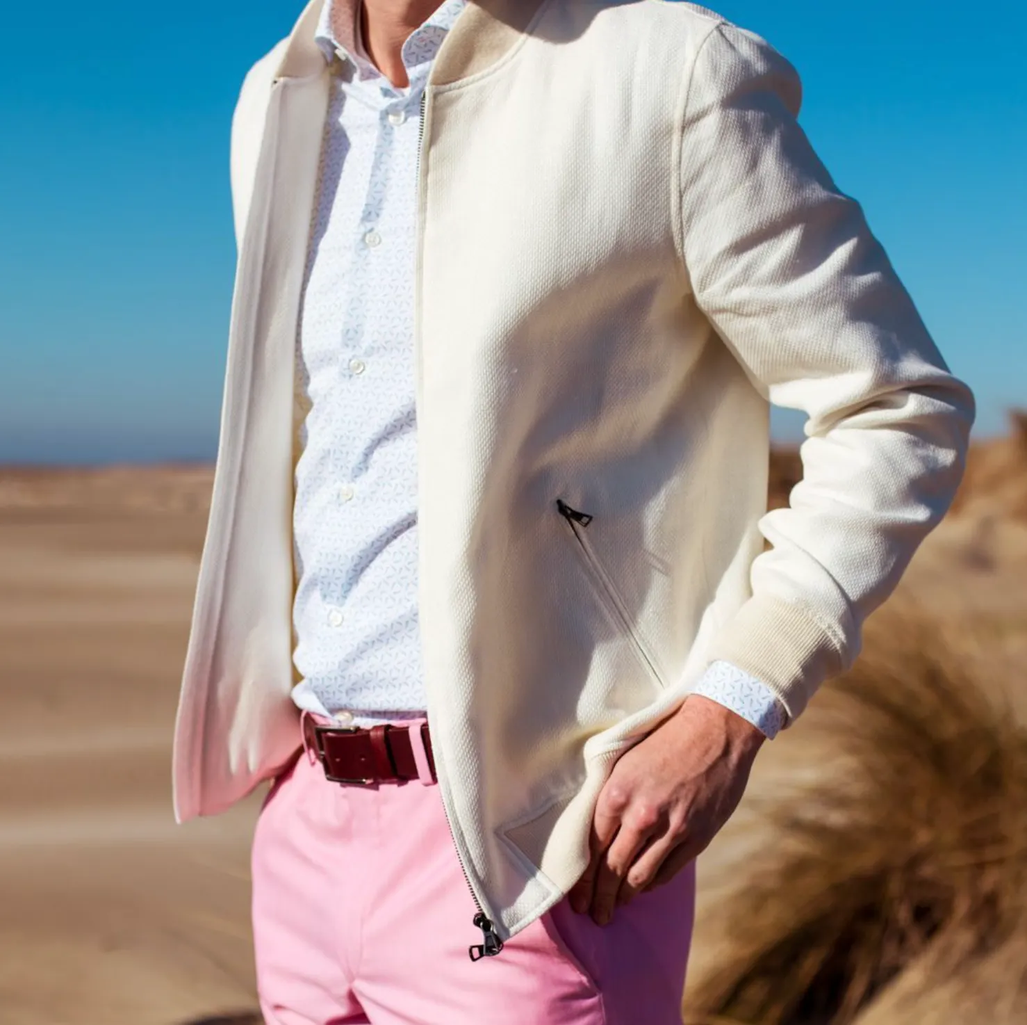 Man wearing a white bomber jacket with pink pants and a burgundy leather belt, showing how a belt completes a smart casual outfit.