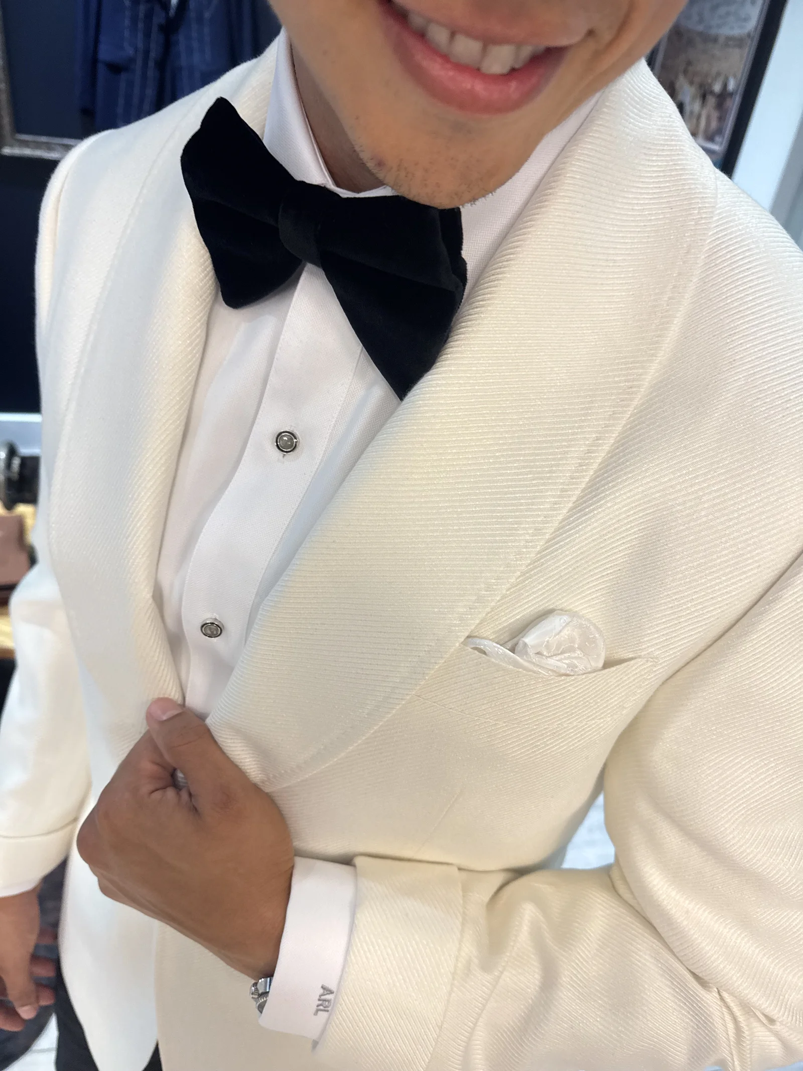 Custom suits: Smiling person in an ivory shawl-collar dinner jacket with a black velvet bow tie, white dress shirt with studs, white pocket square and a monogrammed cuff