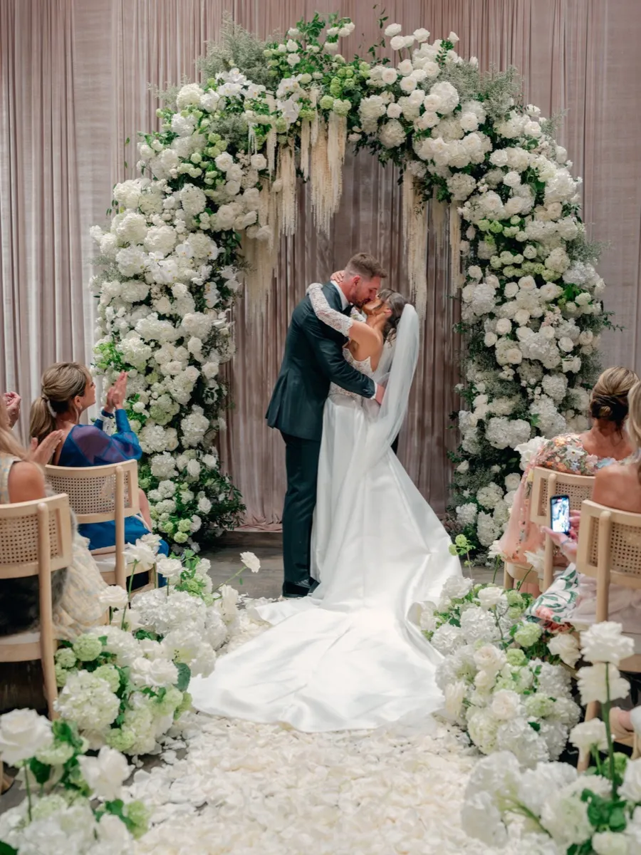 Joey Bosa and Amanda Kassdikian first kiss at the altar framed by floral arch, groom in custom tuxedo by Bespoke By CB
